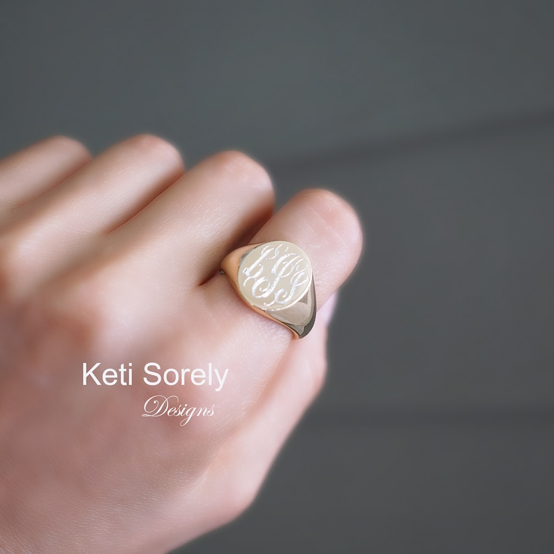 K&ouml;nnte beinhalten: Ein goldener Siegelring mit eingraviertem Monogramm. Der Ring hat eine polierte Oberfl&auml;che und eine abgerundete, ovale Form. Das Monogramm ist in einer Schreibschrift. Der Text "Keti Sorely Designs" befindet sich in der unteren linken Ecke.