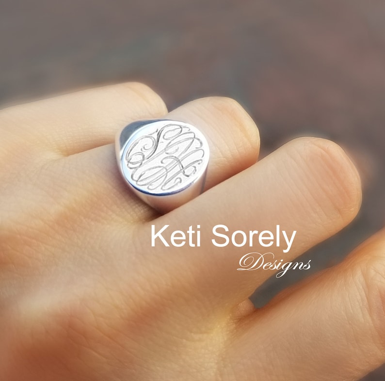 K&ouml;nnte beinhalten: Ein silberner Siegelring mit einem eingravierten Monogramm-Design. Der Ring wird an einem Finger getragen. Der Text "Keti Sorely Designs" ist im Bild zu sehen.