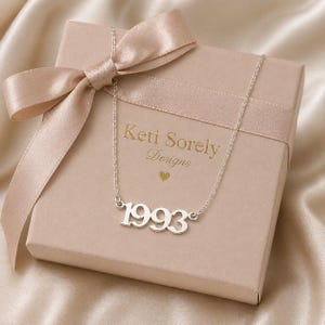 Pode incluir: Um colar prateado com o ano de 1993, repousando sobre uma caixa de presente cor de rosa claro amarrada com uma fita de cetim. A caixa é da marca "Keti Sorely Designs" em letras douradas.