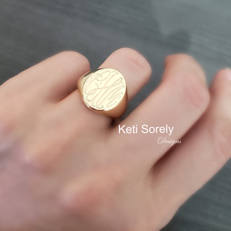 K&ouml;nnte beinhalten: Ein goldener Siegelring mit einem eingravierten Monogramm. Der Ring ist oval und wird am Finger getragen. Der Text "Keti Sorely Designs" ist im Bild sichtbar.
