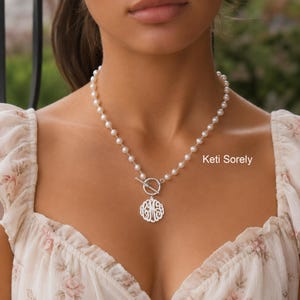 May include: A pearl necklace with a silver monogram pendant. The necklace features a toggle clasp and a decorative, openwork pendant. The text "Keti Sorely" is visible on the right side of the image.