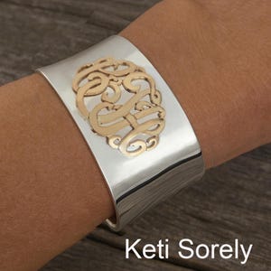 Op de afbeelding: Een brede, zilverkleurige manchetarmband met een goudkleurig monogramontwerp. De armband wordt om de pols gedragen en heeft een gepolijste afwerking. De tekst "Keti Sorely" staat onderaan de afbeelding.