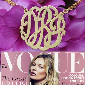 May include: Gold necklace with a large, circular pendant featuring a monogram of the letters "DR" in a decorative, cursive font.