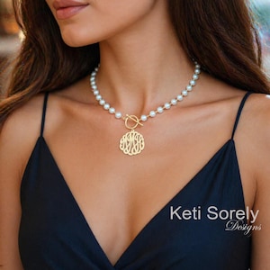 May include: A gold-colored monogram pendant necklace with a pearl strand. The pendant is circular with an intricate, stylized monogram design. The necklace has a toggle clasp and is worn on a person wearing a black dress.