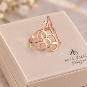 May include: A rose gold monogram ring featuring an intricate, swirling script design. The ring is displayed on a light-colored square box that features the black printed text Keti Sorel Designs and a stylized KS logo on the top surface.