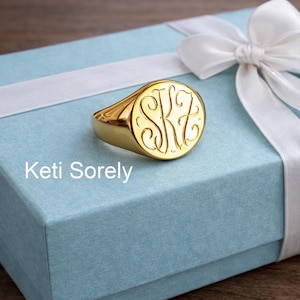 Könnte beinhalten: Ein goldener Siegelring mit eingravierten Initialen, der auf einer hellblauen Geschenkbox mit weißem Band liegt. Die Initialen des Rings sind in einer eleganten, geschwungenen Schriftart gehalten. Der Text "Keti Sorely" ist auf der Box zu sehen.