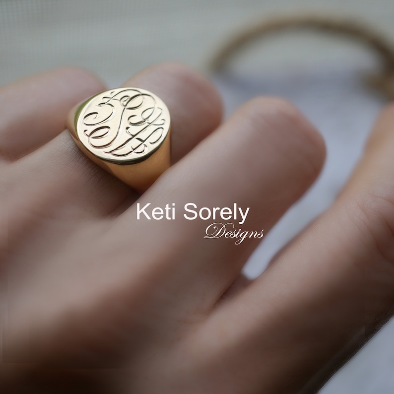 K&ouml;nnte beinhalten: Ein goldener Siegelring mit einem eingravierten Monogramm. Der Ring hat eine polierte Oberfl&auml;che und eine runde Form. Der Text "Keti Sorely Designs" ist im Bild zu sehen.