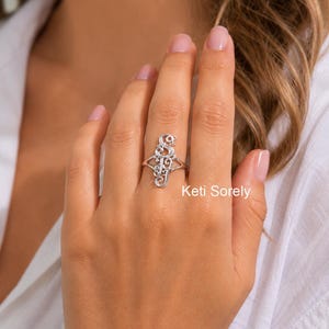 May include: A silver-coloured ring featuring a vertical, ornate, script-style monogram design with interconnected letters. The metal band splits into two thin segments as it connects to the decorative centrepiece, which displays a polished, reflective finish.