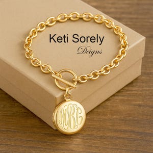 Könnte beinhalten: Goldkette Armband mit einem Schiebeverschluss und einem runden Anhänger mit einem Monogramm. Das Armband liegt auf einer braunen Oberfläche.