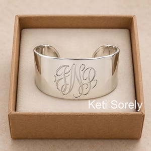 May include: A polished silver cuff bracelet featuring an engraved script monogram of the letters FNP in the centre. The bracelet is presented inside a square cardboard box with a light-coloured interior lining. The text Keti Sorely is visible.