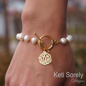 May include: A gold bracelet with a toggle clasp and a circular monogram charm. The bracelet is adorned with white, irregularly shaped pearls. The brand name "Keti Sorely Designs" is visible in the image.