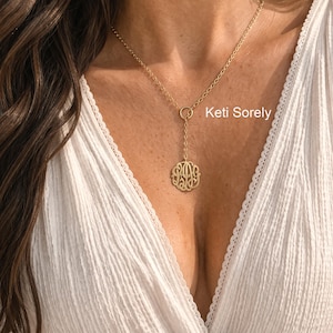 May include: A gold monogram necklace with a fine chain and a circular pendant. The pendant has an intricate, stylized design. The necklace is shown against a white background, with the text "Keti Sorely" visible.