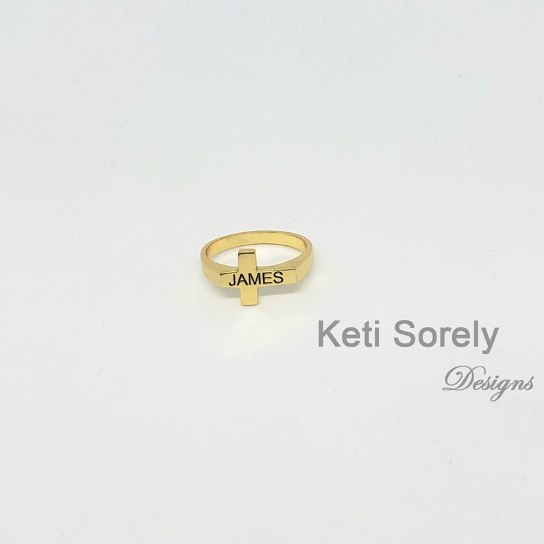 Puede incluir: Anillo dorado con un dise&ntilde;o de cruz y el nombre "JAMES" grabado en la banda.