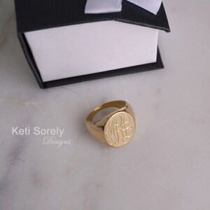 K&ouml;nnte beinhalten: Ein goldener Siegelring mit eingravierten Initialen. Der Ring ist rund mit einer flachen, ovalen Oberfl&auml;che. Die Initialen sind in einer Schreibschrift. Der Ring liegt auf einer Marmoroberfl&auml;che neben einer schwarzen Schachtel mit wei&szlig;em Band.