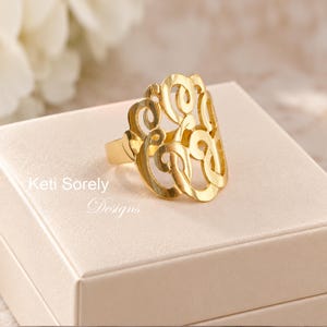May include: A gold-toned ring featuring an intricate, openwork monogram design with swirling, interconnected letters. The ring is displayed on a smooth, light-colored surface with the text Keti Sorely Designs visible nearby.