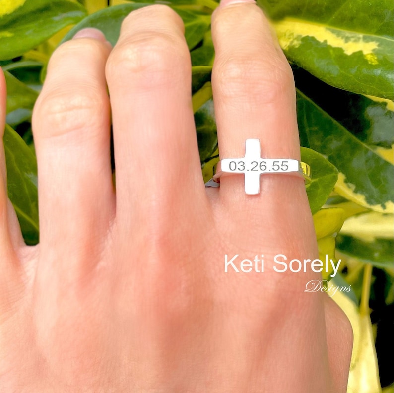 Puede incluir: Un anillo de plata con forma de cruz con la fecha 03.26.55 grabada. El anillo se lleva en un dedo.
