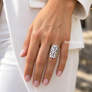 May include: A silver-toned metal ring featuring an intricate, cut-out monogram design with swirling, interconnected letters. The ring has a wide, band-style silhouette that covers a significant portion of the finger. The text Keti Sorely is visible.