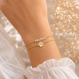 Puede incluir: Dos delicadas pulseras doradas apiladas en una muñeca. La pulsera superior deletrea "Andrea" en cursiva. La pulsera inferior presenta un dije de huella de pata. Las pulseras están en una muñeca con una manga blanca con volantes. El texto "Keti Sorely Designs" está en el fondo.