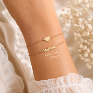 Puede incluir: Dos pulseras doradas se lucen en una muñeca. La pulsera superior tiene un dije de corazón. La pulsera inferior deletrea el nombre "Andrea" en cursiva. También se ve el texto "Keti Sorely Designs".