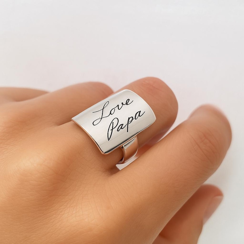 Puede incluir: Anillo de plata con una banda rectangular grabada con las palabras "Love Papa" en cursiva.