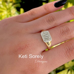 Peut inclure: Une bague signet en or avec un dessus rectangulaire orné d'un monogramme gravé. La bague est portée au doigt.