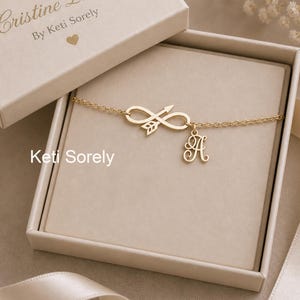 May include: A gold-colored necklace with an infinity symbol and arrow charm, along with a letter "A" pendant, presented in a beige jewelry box. The box lid is open, revealing the necklace. The text "Keti Sorely" is visible.