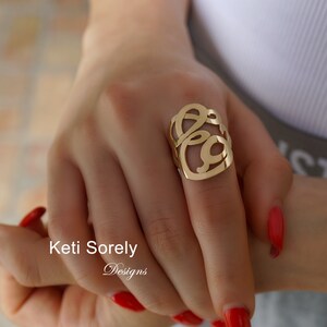 May include: A gold ring with a unique, openwork design featuring a stylised double letter. The ring is worn on a finger with red nail polish.