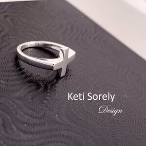 Puede incluir: Un anillo de plata con un dise&ntilde;o de cruz simple. El anillo est&aacute; sobre un fondo negro con el texto "Ketti Sorely Design".