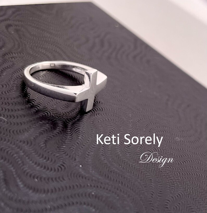 Puede incluir: Un anillo de plata con un dise&ntilde;o de cruz simple. El anillo est&aacute; sobre un fondo negro con el texto "Ketti Sorely Design".