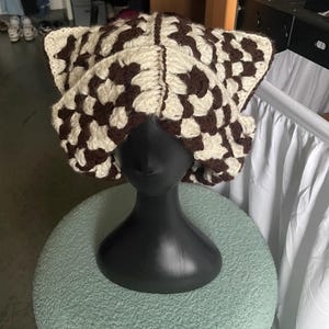 May include: A crochet cat ear hat featuring a checkered pattern in dark brown and cream yarn. The hat is constructed from square motifs joined together, creating a textured surface with pointed ear-like shapes at the top corners of the headwear.