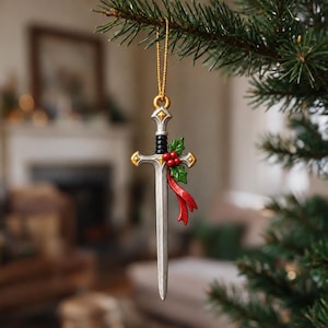 May include: A silver-toned sword ornament hanging from a gold string. The sword features a black grip, a crossguard with gold accents, and a decorative cluster of green holly leaves with red berries and a flowing red ribbon attached to the side.
