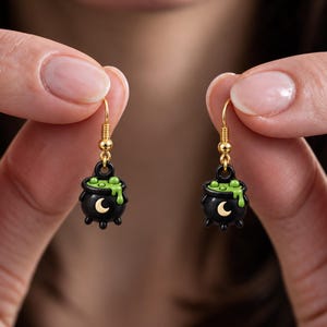 May include: A pair of dangle earrings featuring black cauldron charms with green bubbling liquid spilling over the rim. Each cauldron has a small gold crescent moon symbol on the front and hangs from a gold-toned metal hook wire finding.