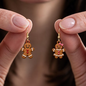 May include: A pair of gold-toned dangle earrings featuring brown gingerbread person charms. One charm has red buttons and white icing details, while the other features a pink bow, pink buttons, and white icing trim on the arms and skirt.