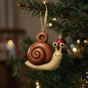 May include: A snail-shaped hanging ornament featuring a dark brown spiral shell and a light beige body with two black eyes and a small smile. The ornament is suspended by a thin gold cord attached to a loop at the top of the snail's shell.