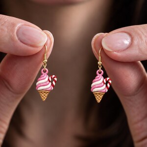 May include: A pair of dangle earrings featuring a miniature ice cream cone charm with pink and white swirled soft serve, a tan waffle cone base, and a small red and white striped candy cane accent, suspended from gold-toned metal hooks.