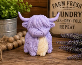 May include: A lavender and cream-colored Highland cow figurine featuring long, shaggy fur texture, curved horns, and a prominent fringe covering the eyes. The figure is sculpted with a solid, opaque finish and displays a seated, forward-facing posture.