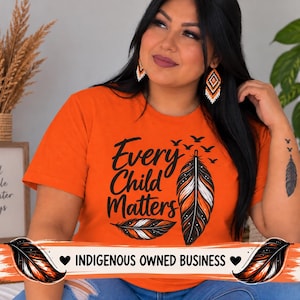 May include: An orange t-shirt featuring the black text Every Child Matters, accompanied by graphic illustrations of two feathers and a flock of birds. The design includes orange, white, and black color accents within the feather graphics.
