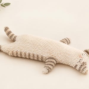 Puede incluir: Un juguete de gato tejido en color beige claro con patrones de rayas marrones en la cola, las patas y las orejas. El gato se muestra en una posición plana y extendida con rasgos faciales bordados, incluyendo una nariz rosa y ojos oscuros.