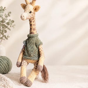 Pode incluir: Um brinquedo de girafa tricotado à mão com pescoço comprido, manchas castanhas num corpo creme e cascos castanhos escuros. A girafa usa uma camisola de malha verde sálvia de manga curta com gola e bainha caneladas e tem uma cauda castanha.