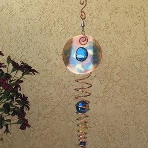 May include: Copper wind chime with a spiral design. The chime features a copper disc with a blue glass stone, a spiral of copper wire, and several colorful glass beads. The wind chime hangs from a black hook.
