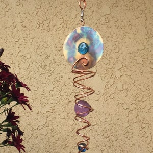 May include: A copper wind chime with a spiral design and three glass beads. The top bead is purple, the middle bead is blue, and the bottom bead is blue with a white swirl. The chime is suspended from a copper hook.