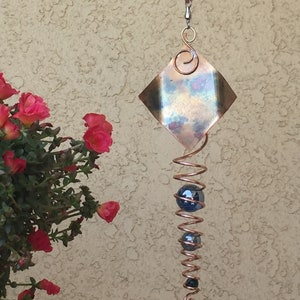 May include: Copper wind chime with a diamond-shaped top and a spiral design. The wind chime features three blue glass spheres and a decorative swirl at the top. The background is a textured beige wall, with red flowers on the left.