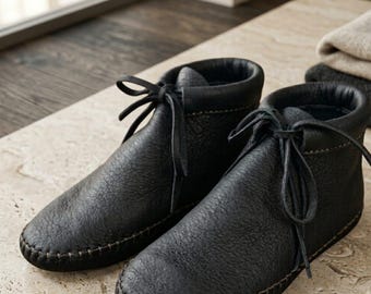 May include: A pair of black leather ankle boots featuring a textured grain finish, a rounded toe, and a moccasin-style construction with visible brown stitching along the sole. The boots include thin black leather laces tied in a bow at the front.