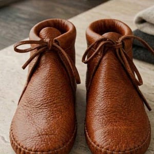May include: A pair of ankle-high boots crafted from textured brown leather. These boots feature a rounded toe, a visible stitched seam along the base, and a matching leather lace-up closure tied in a bow at the front of each boot collar.