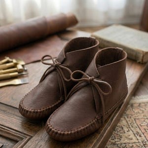 Könnte beinhalten: Ein Paar knöchelhohe Mokassin-Stiefel aus braunem Leder mit weicher, abgerundeter Zehenpartie, sichtbaren Steppnähten entlang der Sohlenkante und dünnen Lederschnürsenkeln, die vorne am Schaft jedes Stiefels zu einer Schleife gebunden sind.