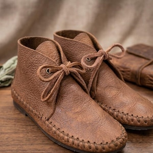 Könnte beinhalten: Ein Paar knöchelhohe Stiefel aus strukturiertem braunem Leder. Die Stiefel verfügen über eine Mokassin-Konstruktion mit sichtbaren Ziernähten entlang der Sohlenkante sowie eine Schnürung mit zwei Ösen und passenden Lederbändern.