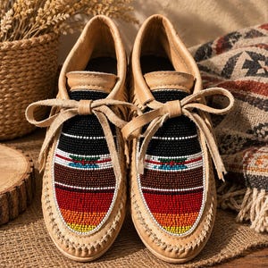 May include: A pair of tan leather moccasins featuring fringe detailing and a lace-up front. The vamp is decorated with a multi-colored beaded pattern in horizontal stripes of black, white, teal, brown, red, orange, and yellow beads.