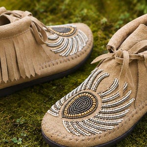 May include: A pair of tan suede moccasins featuring fringe detailing around the ankle and a decorative beaded pattern on the top. The beadwork consists of concentric teardrop shapes in black, gold, and white, with thin laces tied at the front.