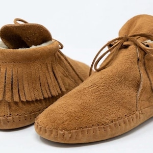 May include: A pair of tan suede moccasins featuring a soft, light-coloured interior lining, a fringe detail around the ankle collar, and thin leather laces tied at the front. The shoes have visible whipstitching along the outer sole edge.