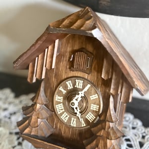 May include: A wooden cuckoo clock with a brown finish. The clock has a traditional house shape with a sloped roof and a round clock face with Roman numerals. The clock hands are white. The clock is sitting on a white lace surface.